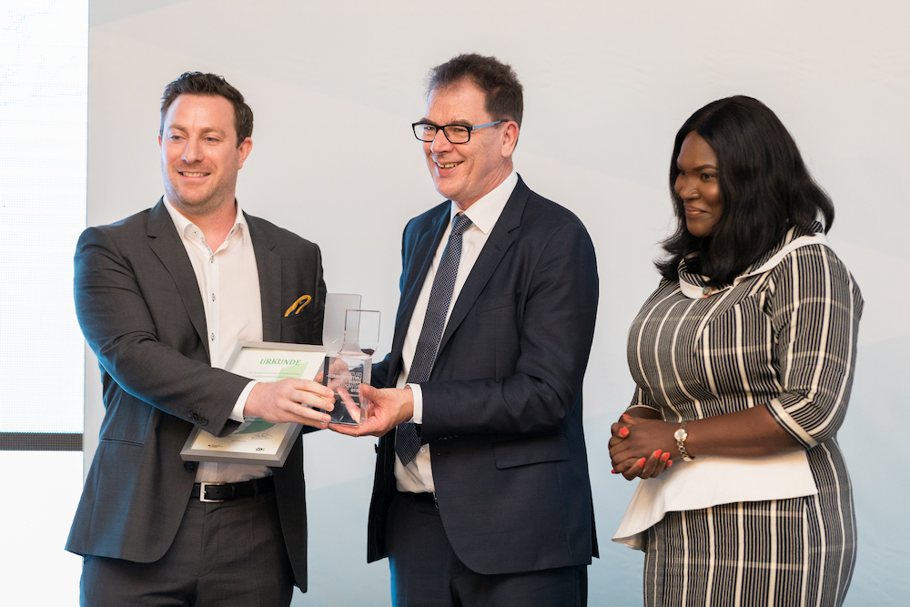 Giám đốc điều hành ecoligo Martin Baart nhận giải thưởng từ Bộ trưởng Hợp tác Kinh tế và Phát triển Liên bang Gerd Müller. Người điều hành là Anita Erskine. © THEGREYSHUTTER_2019